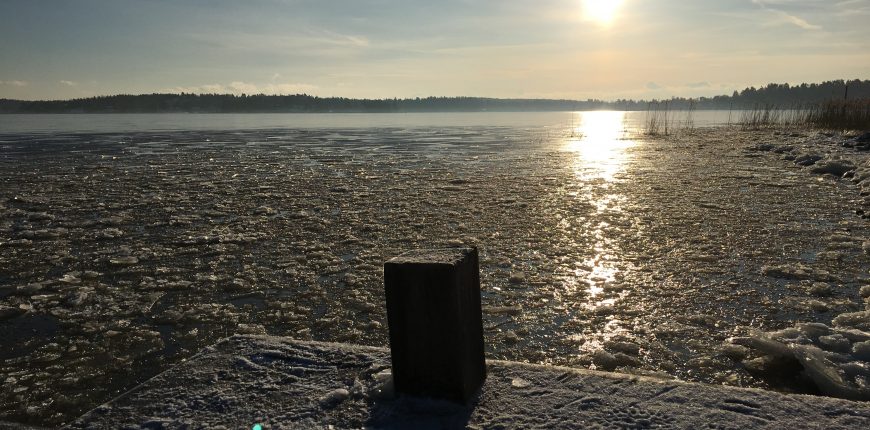 En snöklädd brygga i förmiddagssolen som lyser på isen som ligger i bitar vid strandkanten.