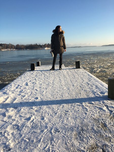 Mörkhårig kvinna på snöklädd brygga. Står med kroppen mot kameran, huvudet vänt mot solen.