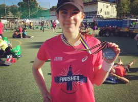 Kvinna i rosa tränings- t-shirt och svart keps håller upp en medalj.