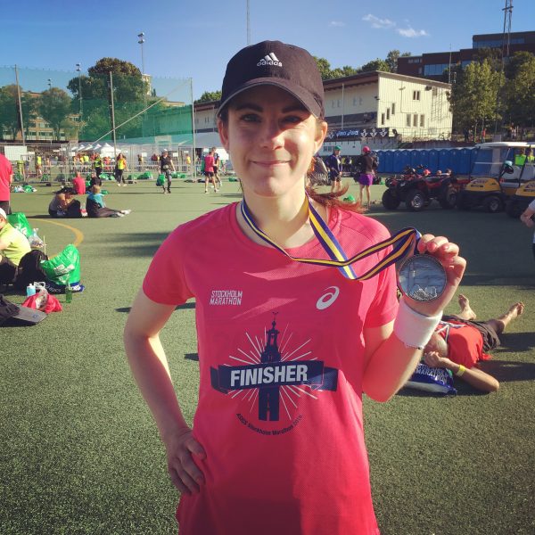 Kvinna i rosa tränings- t-shirt och svart keps håller upp en medalj.